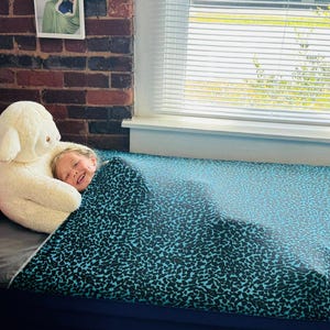 May include: A child smiles while snuggled under a teal and black patterned blanket. A large white stuffed sheep sits next to the child. A red pillow and a brick wall are in the background.