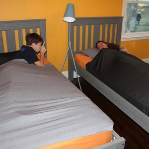 May include: Two twin beds with gray headboards and orange sheets. One bed has a gray weighted blanket, the other has a black weighted blanket. A boy is on one bed, and a girl is on the other. A gray floor lamp stands between the beds.