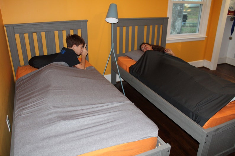 May include: Two twin beds with gray headboards and orange sheets. One bed has a gray weighted blanket, the other has a black weighted blanket. A boy is on one bed, and a girl is on the other. A gray floor lamp stands between the beds.