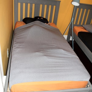 May include: A gray twin-sized bed frame with a gray mattress and orange fitted sheet. The bed is made with a gray blanket and a person is lying under the blanket.