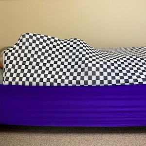 May include: A bed covered with a black and white checkered blanket. The bed has a purple bed skirt and a gray mattress. The bed is against a beige wall and sits on a carpeted floor.