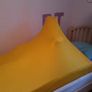 May include: A bright yellow sensory sock stretched over a bed. The fabric is pulled taut, creating a tent-like shape. A blue pillow and a wooden bed frame are visible in the background. The sock appears to be made of a stretchy material.