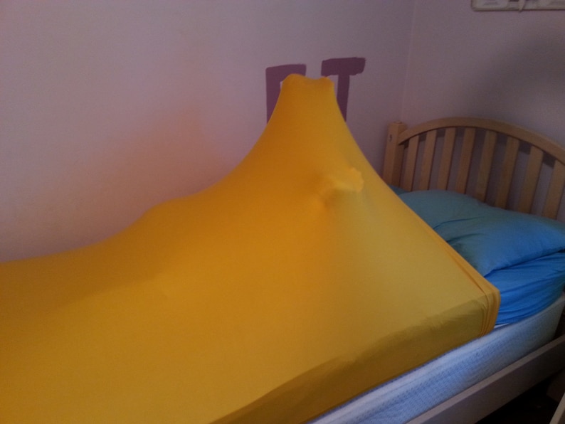 May include: A bright yellow sensory sock stretched over a bed. The fabric is pulled taut, creating a tent-like shape. A blue pillow and a wooden bed frame are visible in the background. The sock appears to be made of a stretchy material.