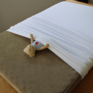 May include: A bed with a brown mattress and white sheets. A small teddy bear wearing a blue and white striped shirt with a red heart is tucked under the sheets. The bed is on a wooden frame.