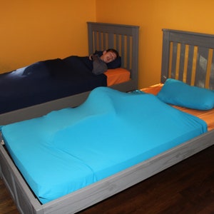 May include: Two beds with gray frames and orange sheets. One bed has a navy blue sleep sack, and the other has a turquoise sleep sack. A turquoise pillow rests on the second bed. The room has yellow walls.