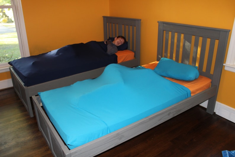 May include: Two beds with gray frames and orange sheets. One bed has a navy blue sleep sack, and the other has a turquoise sleep sack. A turquoise pillow rests on the second bed. The room has yellow walls.