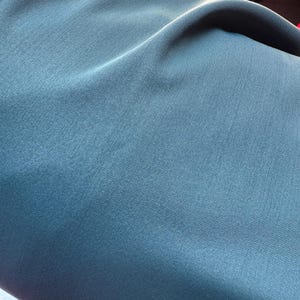 May include: Close-up of a teal fabric with a subtle ribbed texture. The fabric is folded, creating soft shadows and highlights across its surface. The color is a muted, cool tone, suggesting a smooth, possibly synthetic material.