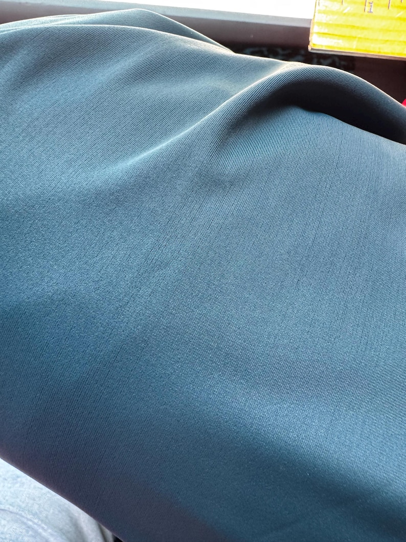 May include: Close-up of a teal fabric with a subtle ribbed texture. The fabric is folded, creating soft shadows and highlights across its surface. The color is a muted, cool tone, suggesting a smooth, possibly synthetic material.