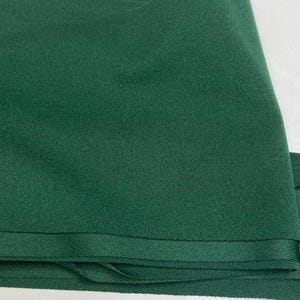 May include: Close-up of a folded piece of dark green fabric. The fabric has a smooth texture and a slight sheen. The edges are neatly finished with a sewn hem. The fabric appears to be a solid color, with no visible patterns or designs.