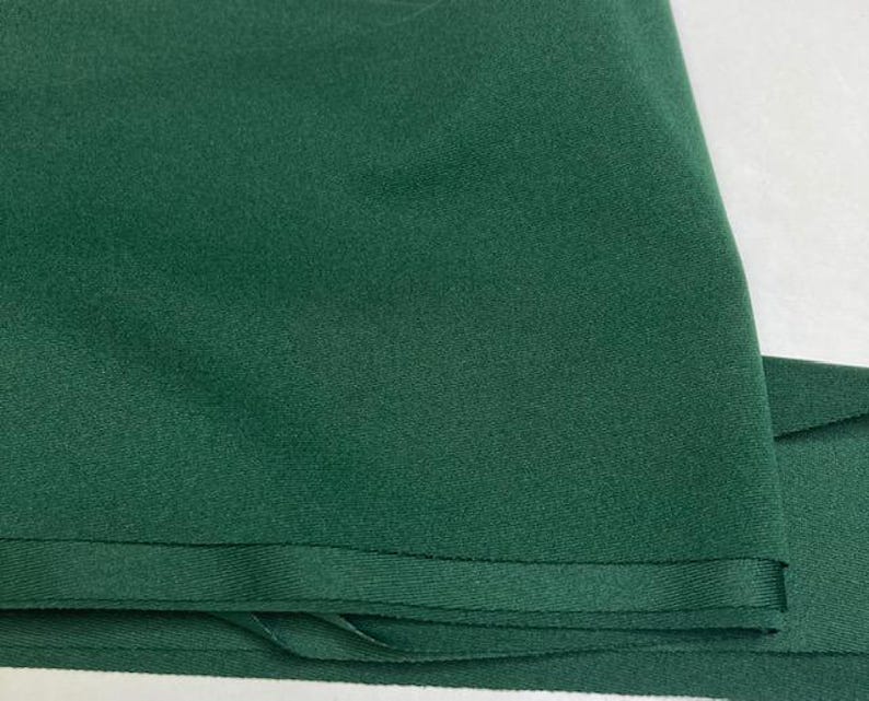 May include: Close-up of a folded piece of dark green fabric. The fabric has a smooth texture and a slight sheen. The edges are neatly finished with a sewn hem. The fabric appears to be a solid color, with no visible patterns or designs.