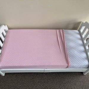 May include: A white wooden toddler bed with a pink fitted sheet and a patterned sheet. The bed frame has a curved headboard and footboard. The pink sheet has a subtle dotted pattern.