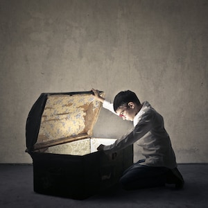 May include: A young person wearing glasses is kneeling and looking inside an old wooden treasure chest. The chest is open and glowing from within.