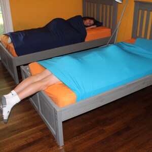 May include: Two beds with gray frames and orange bedding. One bed has a person inside a navy blue sleep sack. The other bed has a person with legs exposed, covered by a turquoise sleep sack. The room has yellow walls and a wooden floor.