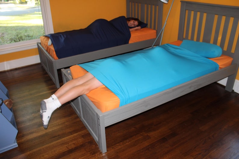 May include: Two beds with gray frames and orange bedding. One bed has a person inside a navy blue sleep sack. The other bed has a person with legs exposed, covered by a turquoise sleep sack. The room has yellow walls and a wooden floor.