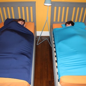 May include: Two beds with gray headboards and orange sheets. One bed has a navy blue sleep sack, the other a turquoise sleep sack. A blue lamp stands between the beds. The room has yellow walls and a dark wood floor.