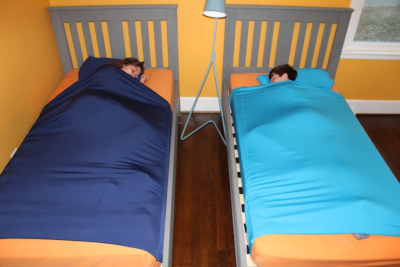 May include: Two beds with gray headboards and orange sheets. One bed has a navy blue sleep sack, the other a turquoise sleep sack. A blue lamp stands between the beds. The room has yellow walls and a dark wood floor.