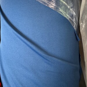 May include: A roll of blue fabric, tightly wrapped in clear plastic. The fabric has a ribbed texture, with vertical lines running down its surface. The color is a medium shade of blue, and the fabric appears to be soft and flexible.