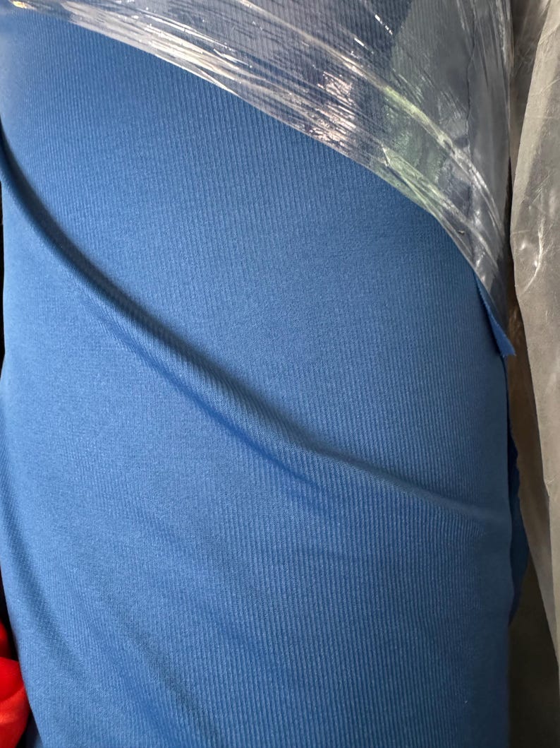 May include: A roll of blue fabric, tightly wrapped in clear plastic. The fabric has a ribbed texture, with vertical lines running down its surface. The color is a medium shade of blue, and the fabric appears to be soft and flexible.