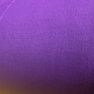 May include: Close-up of a roll of purple fabric. The fabric has a ribbed texture with vertical lines. The edges of the fabric are visible, showing the layers and the cut edges. The color is a vibrant, solid purple.