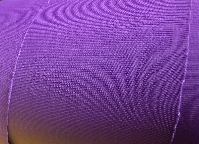 May include: Close-up of a roll of purple fabric. The fabric has a ribbed texture with vertical lines. The edges of the fabric are visible, showing the layers and the cut edges. The color is a vibrant, solid purple.