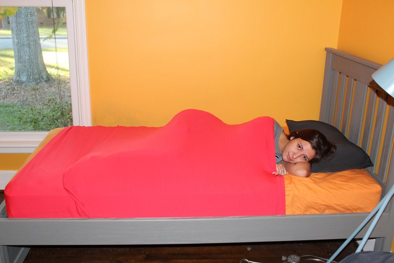 May include: A red sensory compression bed sheet covers a bed with a gray frame. The bed has an orange sheet and a black pillow. The room has yellow walls and a window with a view of a tree.