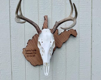May include: A white deer skull with antlers mounted on a wooden plaque shaped like the state of West Virginia. The wood features dark, burnt edges and is engraved with the text: Lainey Leyda, Putnam Co., 11/11/25, positioned on the left side.