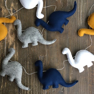 May include: A garland of felt dinosaurs in various colors, including gray, white, blue, and yellow. The dinosaurs are strung together with white thread and are hanging against a wood background.