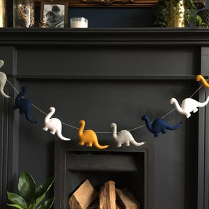 May include: A felt dinosaur garland with blue, white, gray, and yellow dinosaurs hanging from a string. The garland is hanging above a fireplace.