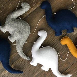 May include: A garland of five felt dinosaur shapes, including gray, white, navy blue, and yellow. The dinosaurs are strung together with white thread.