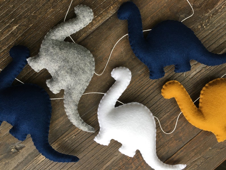 May include: A garland of five felt dinosaur shapes, including gray, white, navy blue, and yellow. The dinosaurs are strung together with white thread.