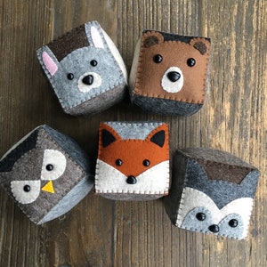 Peut inclure: Cinq cubes en feutre représentant des animaux avec des visages brodés. Les cubes présentent un renard, un hibou, un ours et deux loups différents. Les cubes sont tous gris, bruns et blancs.