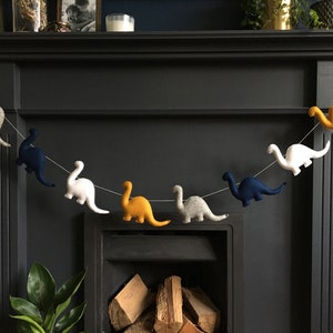 May include: A garland of felt dinosaurs in various colors, including yellow, white, blue, and gray, hangs above a fireplace. The dinosaurs are strung together with white thread.