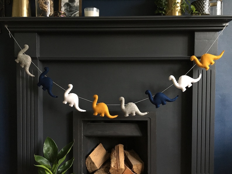 May include: A garland of felt dinosaurs in various colors, including yellow, white, blue, and gray, hangs above a fireplace. The dinosaurs are strung together with white thread.