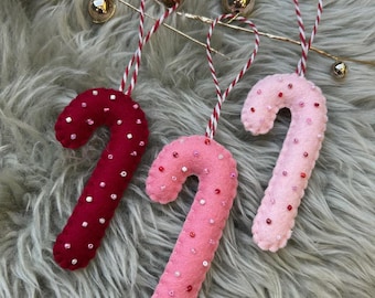 Puede incluir: Tres adornos de fieltro con forma de bastón de caramelo en tonos rojos y rosas, adornados con pequeñas cuentas de colores. Cada bastón tiene una cuerda a rayas rojas y blancas para colgar. Decoración festiva navideña.