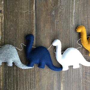 May include: A garland of felt dinosaurs in gray, navy blue, white, and yellow. The dinosaurs are strung together with white thread.