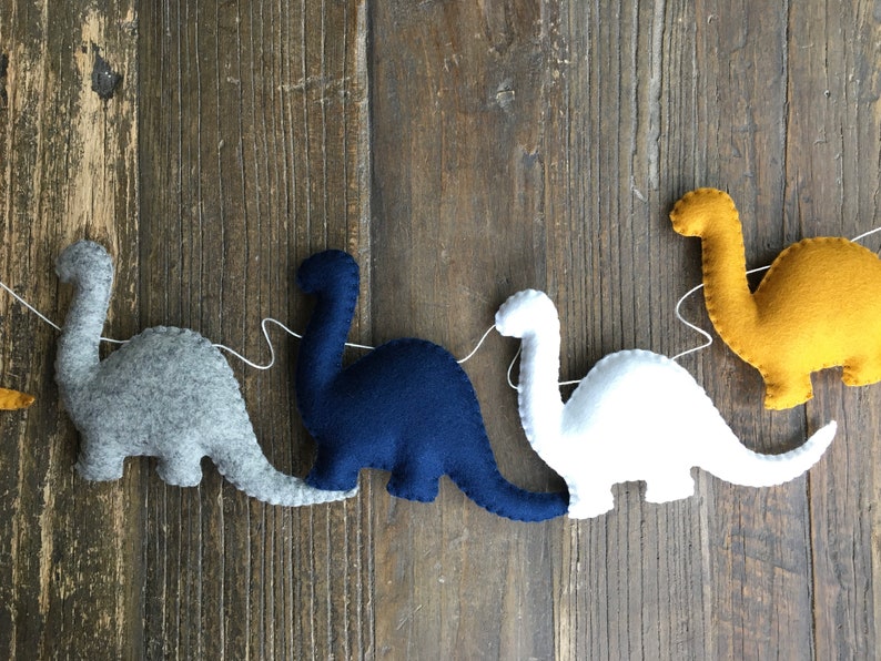 May include: A garland of felt dinosaurs in gray, navy blue, white, and yellow. The dinosaurs are strung together with white thread.