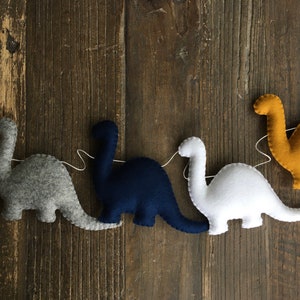 May include: A garland of five felt dinosaurs in different colors, including gray, navy blue, white, and yellow. The dinosaurs are strung together with white thread.