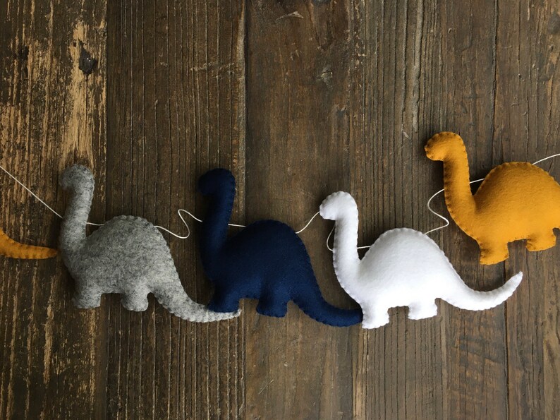 May include: A garland of five felt dinosaurs in different colors, including gray, navy blue, white, and yellow. The dinosaurs are strung together with white thread.