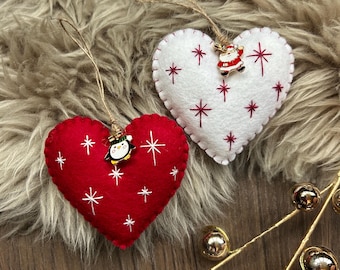 Puede incluir: Dos adornos navideños en forma de corazón. Uno es rojo con bordados de estrellas blancas y un dije de pingüino. El otro es blanco con bordados de estrellas rojas y un dije de Papá Noel. Ambos tienen lazos de cuerda para colgar. Decoración festiva.