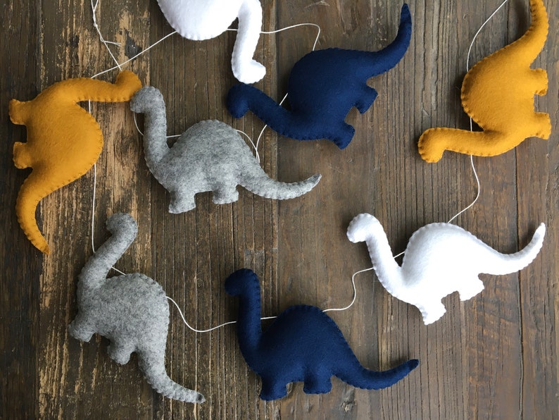 May include: A garland of felt dinosaurs in various colors, including gray, white, blue, and yellow. The dinosaurs are strung together with white thread and are hanging against a wood background.