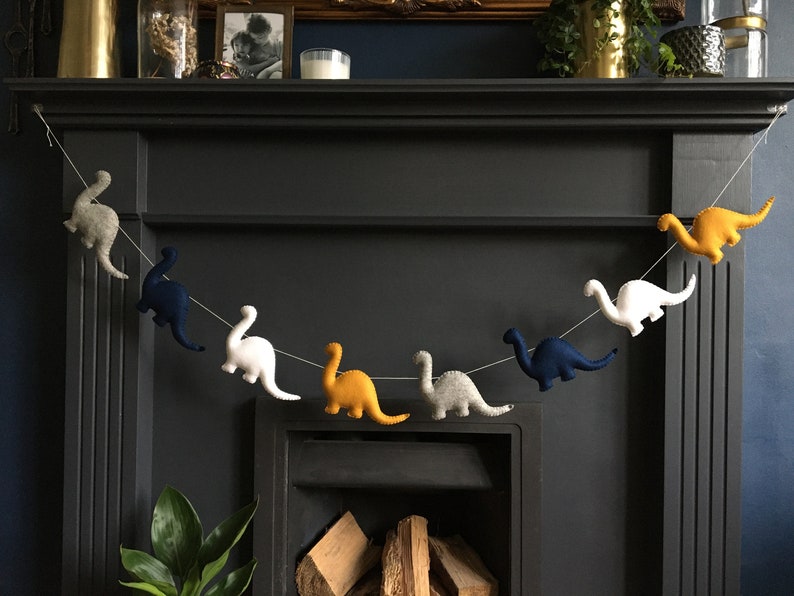 May include: A felt dinosaur garland with blue, white, gray, and yellow dinosaurs hanging from a string. The garland is hanging above a fireplace.