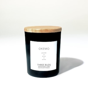 May include: Black candle jar with a wooden lid. The label reads "OKEMO" and lists the scent notes "powder, pines, winter". The label also reads "THREE BUDS APOTHECARY" and "HAND POURED IN VERMONT".