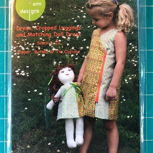 May include: A sewing pattern package from Olive Ann Designs, titled "Peaceout." The package shows a young girl wearing a dress and holding a doll in a matching dress. The pattern includes options for dresses, cropped leggings, and doll dresses, sizes 2-10.