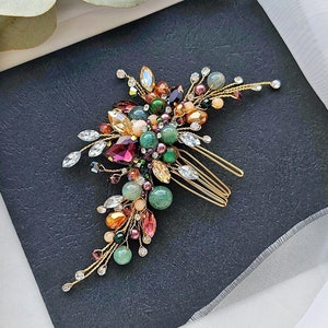 May include: A gold hair comb decorated with a variety of colourful gemstones and crystals. The design includes a cluster of green, pink, orange, and purple stones, with clear crystals. The comb is set against a dark background with white fabric.