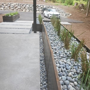 May include: A long, narrow planter made of rusted metal is filled with grey river rocks and green plants. The planter is positioned next to a concrete walkway.