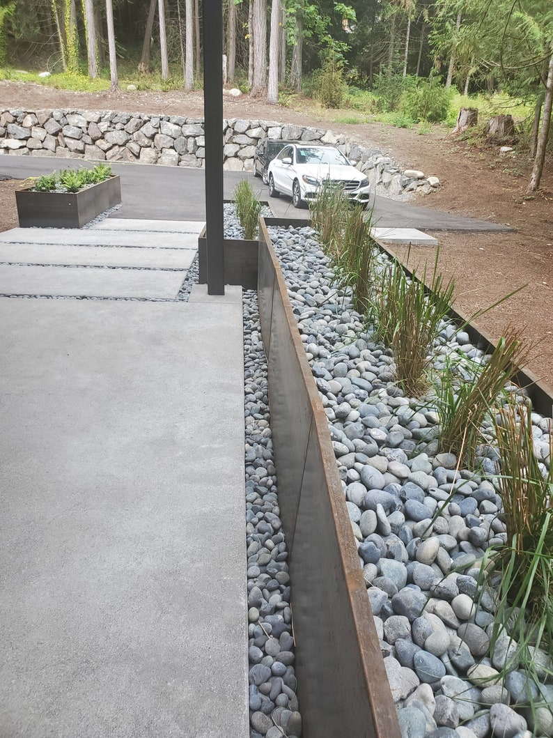 May include: A long, narrow planter made of rusted metal is filled with grey river rocks and green plants. The planter is positioned next to a concrete walkway.