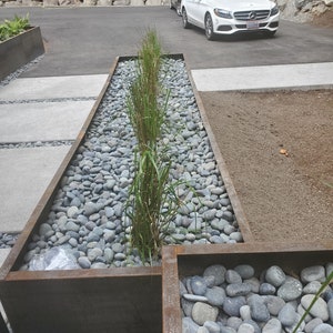 May include: A long, rectangular planter made of rusted metal is filled with grey river rocks and green plants. The planter is set on a concrete patio with a white car parked in the background.