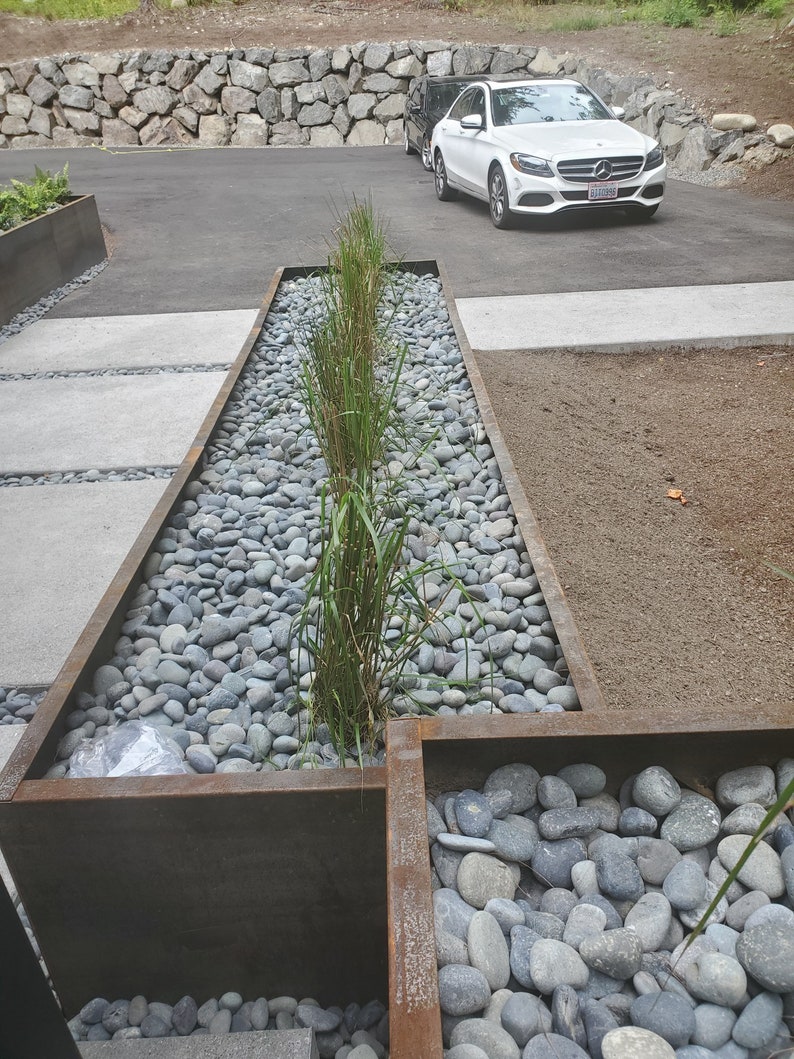 May include: A long, rectangular planter made of rusted metal is filled with grey river rocks and green plants. The planter is set on a concrete patio with a white car parked in the background.