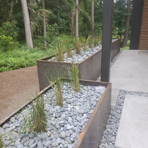 May include: A long, rectangular planter made of rusted metal is filled with grey river rocks and green plants. The planter is placed on a concrete patio with a grey stone path leading to it.