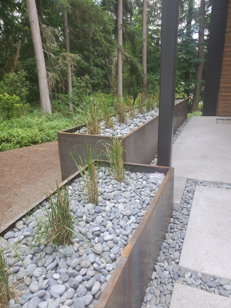May include: A long, rectangular planter made of rusted metal is filled with grey river rocks and green plants. The planter is placed on a concrete patio with a grey stone path leading to it.